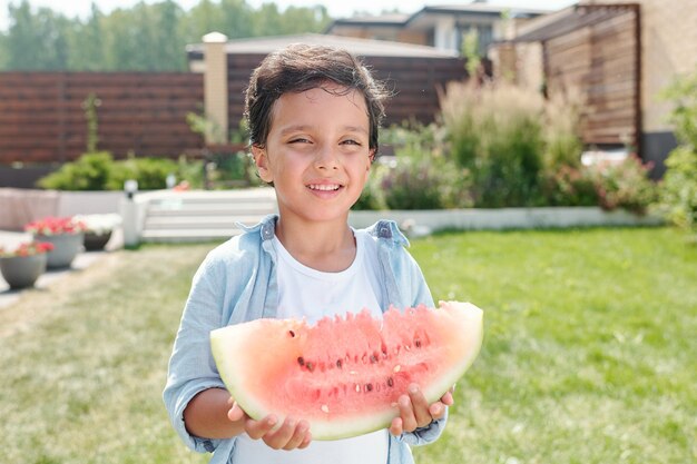 Porträtaufnahme eines fröhlichen kleinen Jungen, der im Hinterhof steht und ein Stück Wassermelone lächelnd hält