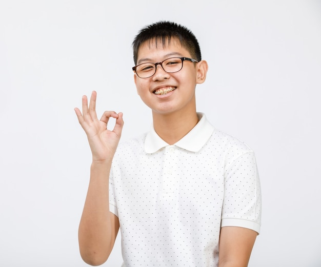 Porträtaufnahme eines hübschen lächelnden jungen Teenagers, der eine Brille trägt und in die Kamera schaut und eine ok Zeichengeste mit den Fingern zeigt, die auf weißem Hintergrund im Studio isoliert sind. Erfolgskonzept