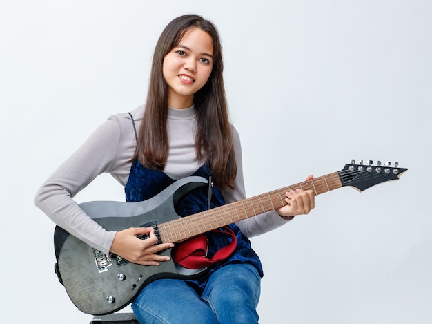 Porträtaufnahme eines süßen lächelnden thailändisch-türkischen Teenagers, der die E-Gitarre spielt. Junior-Gitarrist, der ein Instrument spielt, während er die Kamera einzeln in weißem Hintergrund betrachtet. Konzept des Hobbys