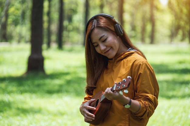 Porträtbild einer schönen asiatischen Frau, die gerne Musik mit Kopfhörern hört, während sie im Park Ukulele spielt