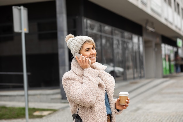 Porträtfrau in Strickmütze und Pelzmantel aus Kunstpelz mit einer Tasse heißem Kaffee in der Hand, die mit dem Handy spricht, Streetstyle, modernes Leben