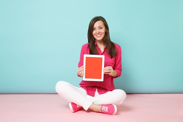 Porträtfrau in voller Länge in rosa Hemdbluse, weiße Hosen, die auf dem Boden sitzen, halten Tablet-PC isoliert auf hellrosa blauer Pastellwand.