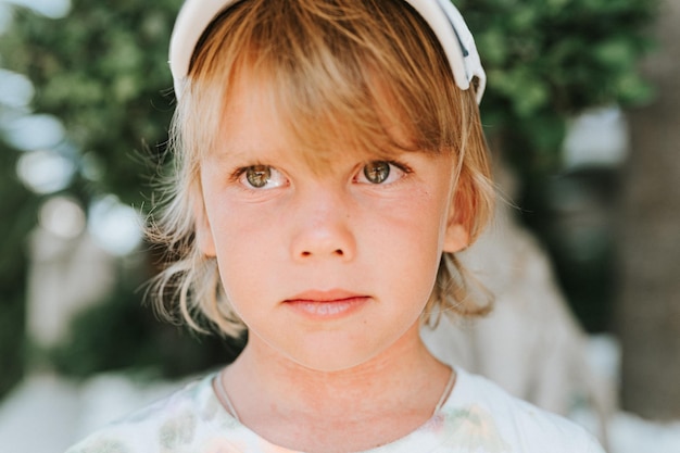 Porträtgesicht eines aufrichtigen, glücklichen kleinen Jungen von fünf Jahren mit problematischer allergischer Haut und langen blonden Haaren und grünen Augen auf dem Hintergrund grüner Pflanzen während einer Sommerferienreise