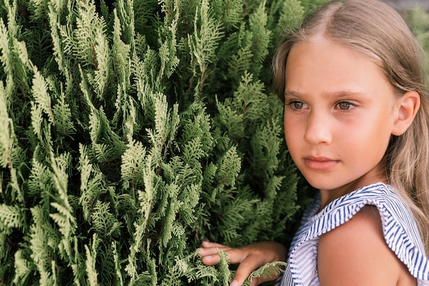 Porträtgesicht eines offenen, glücklichen kleinen Mädchens von acht Jahren mit langen blonden Haaren und grünen Augen auf dem Hintergrund grüner Pflanzen während eines Sommerurlaubsreise-Gen-Z-Konzepts für psychische Gesundheit