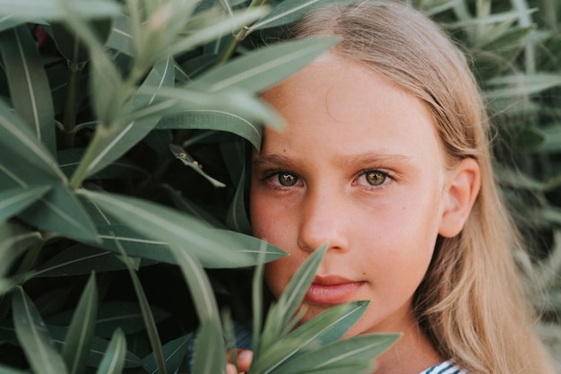 Porträtgesicht eines offenen, glücklichen kleinen Mädchens von acht Jahren mit langen blonden Haaren und grünen Augen auf dem Hintergrund grüner Pflanzen während eines Sommerurlaubsreise-Gen-Z-Konzepts für psychische Gesundheit