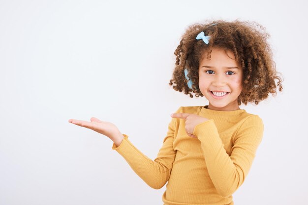 Porträtlächeln und Kind, das auf die Handfläche im Studio zeigt, isoliert auf einem weißen Hintergrund. Gesichts-Hand-Attrappe und glückliche Mädchen-Kind-Werbeshow, Marketing und Präsentation kommerzieller Werbung