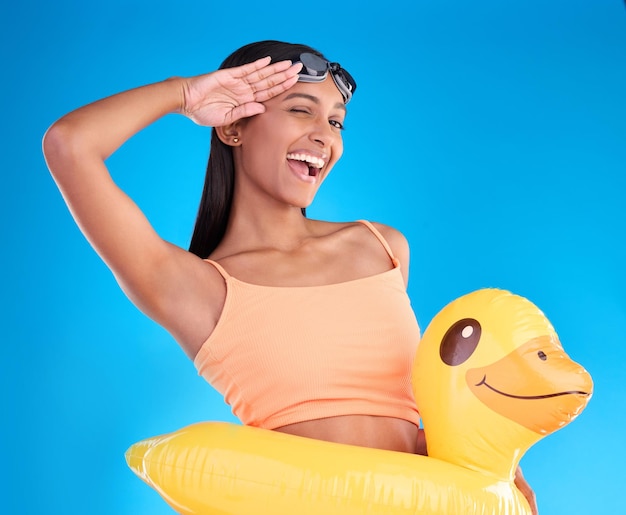 Porträtlächeln und Salutieren mit einer Schwimmerin im Studio auf blauem Hintergrund, die eine Schutzbrille auf dem Kopf trägt. Fröhliche Handbewegung und Schwimmen mit einer attraktiven jungen Frau, die im Sommer gerne schwimmen geht