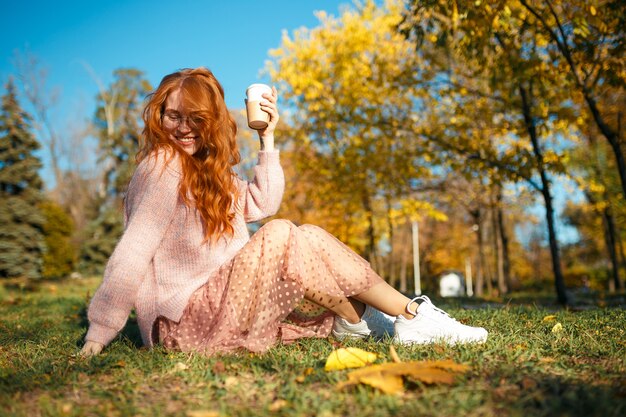 Porträts eines charmanten rothaarigen Mädchens mit Brille und hübschem Gesicht. Mädchen, das im Herbstpark in einem Pullover und in einem Rock der Korallenfarbe aufwirft.