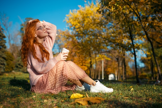 Porträts eines charmanten rothaarigen Mädchens mit Brille und hübschem Gesicht. Mädchen, das im Herbstpark in einem Pullover und in einem Rock der Korallenfarbe aufwirft.