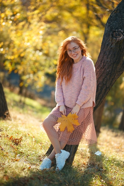 Porträts eines charmanten rothaarigen Mädchens mit einem niedlichen Gesicht. Mädchen, das im Herbstpark in einem Pullover und in einem korallenfarbenen Rock aufwirft. In den Händen eines Mädchens ein gelbes Blatt