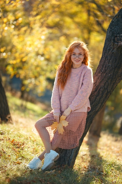 Porträts eines charmanten rothaarigen Mädchens mit einem niedlichen Gesicht. Mädchen, das im Herbstpark in einem Pullover und in einem korallenfarbenen Rock aufwirft. In den Händen eines Mädchens ein gelbes Blatt