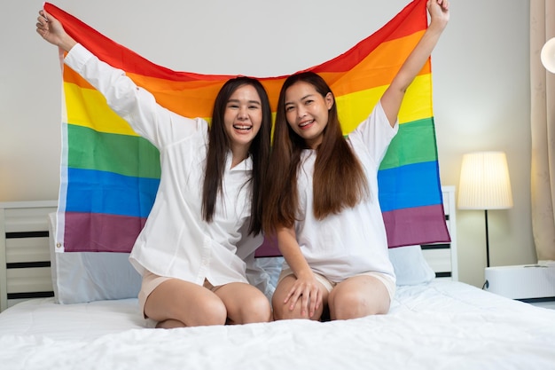 Porträts eines glücklichen asiatischen lesbischen Paares, das mit einer Regenbogenflagge neben dem Bett sitzt