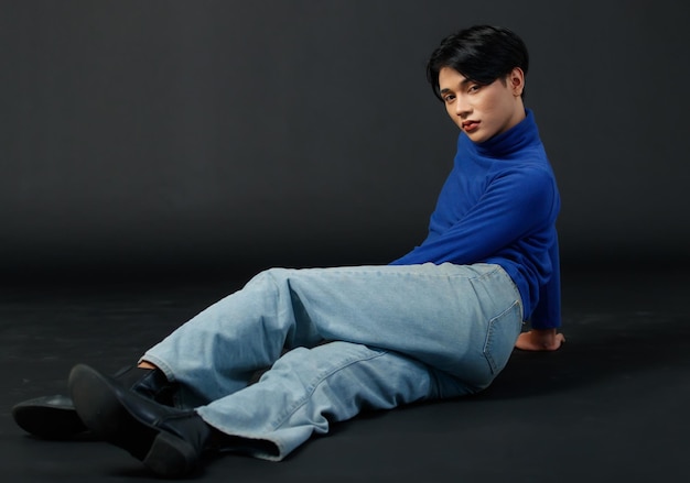 Porträtstudio-Nahaufnahme eines asiatischen jungen sexy Luxus-Glamour schlanken modischen LGBTQ-schwulen männlichen Modells in Rollkragen-Langarmhemd-Jeans-Lederstiefeln, die glattes Haar sitzen und auf schwarzem Hintergrund posieren