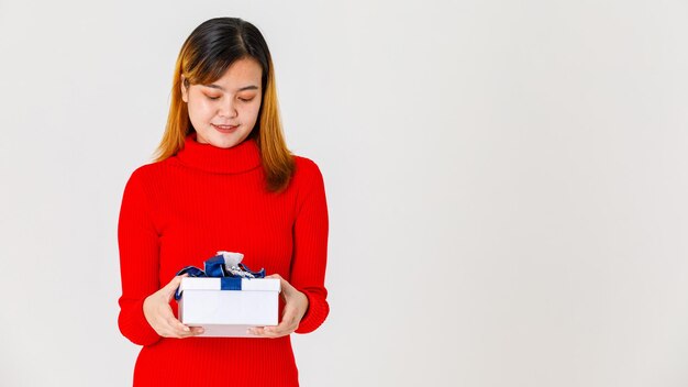 Porträtstudioaufnahme des asiatischen jungen glücklichen weiblichen Geburtstagsmädchens im roten langärmeligen Pulloverkleid, das Geschenkbox mit blauem silbernem Band in den Händen hält, die wichtigen Tag auf weißem Hintergrund feiert.