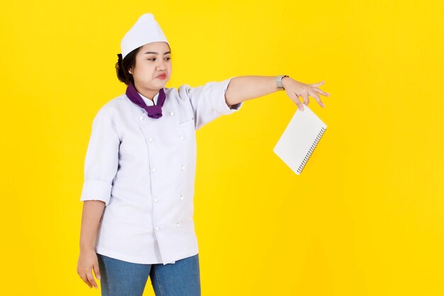 Porträtstudioaufnahme einer asiatischen professionellen Küchenchefin in weißer Kochuniform mit Hut und Schal, die lächelnd hält und neue Rezepte aus dem Notizbuch in den Händen auf gelbem Hintergrund lernt.