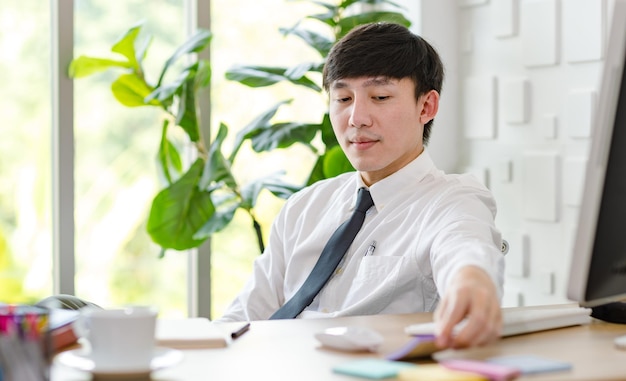 Porträtstudioaufnahme eines asiatischen, professionellen, erfolgreichen männlichen Geschäftsmannangestellten in formellem Hemd mit Krawatte, der am Arbeitstisch mit Computermonitor, Tastatur, Maus und Schreibwaren in die Kamera blickt.