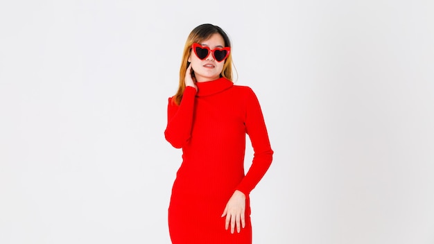 Porträtstudioaufnahme eines asiatischen selbstbewussten weiblichen Modemodells in lässigem modischem, rotem, langärmeligem Rollkragenkleid mit herzförmiger schwarzer Linsensonnenbrille steht sexy posiert auf weißem Hintergrund.