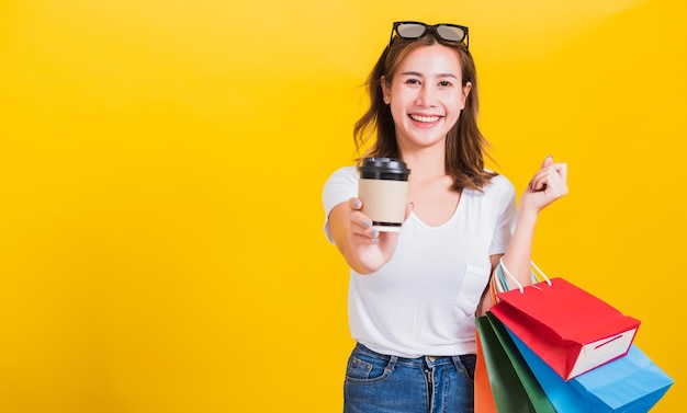 Portrait Asian Thai schöne glückliche junge Frau lächelnd halten Sie Einkaufstaschen mehrfarbig und nehmen Sie Kaffeetasse weg, die sie zur Kamera schaut, Studioaufnahme isoliert auf gelbem Hintergrund, mit Kopierraum