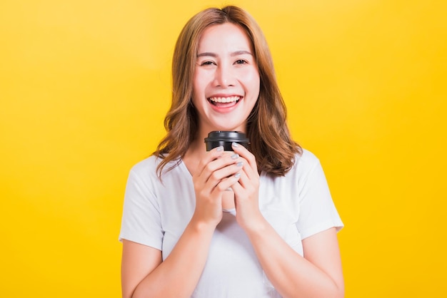 Portrait Asian Thai schöne glückliche junge Frau tragen weißes T-Shirt stehend lächelnd Holding Kaffee Pappbecher zum Mitnehmen, Studioaufnahme isoliert auf gelbem Hintergrund, mit Kopierraum