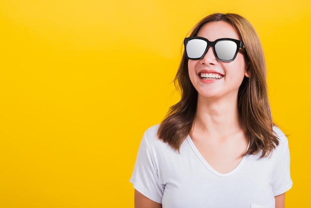 Portrait Asian Thai schöne junge Frau glücklich lächelnde weiße Zähne mit Sonnenbrille und Blick auf den Seitenraum, Studioaufnahme isoliert auf gelbem Hintergrund, mit Kopierraum