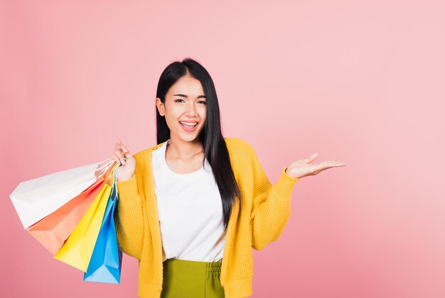 Portrait Asiatin glücklich schöne junge Frau Shopper lächelnd stehend aufgeregt halten Einkaufstaschen mehrfarbig im Sommer zeigen etwas Handfläche, um Raum zu kopieren, Studioaufnahme isoliert auf rosa Hintergrund