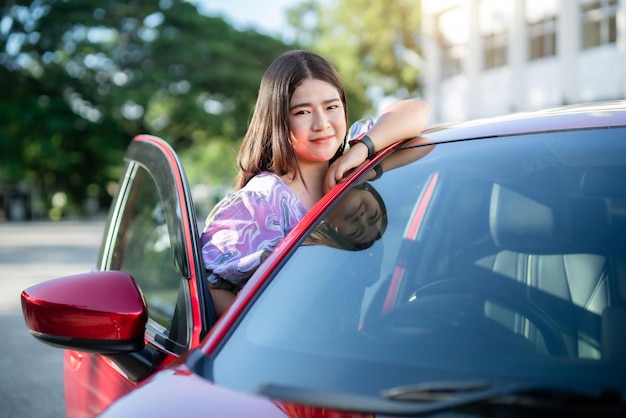 Portrait asiatische Frau Tragen Sie ein lila Kleid mit Smartwatch in der Autotür Modernes rotes Auto im Stadtpark im Freien