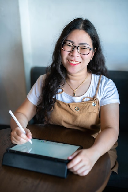 Portrait asiatische freiberufliche Geschäftsfrau mit Brille stilvolle Hipste Zeichnen oder Notizen auf einem digitalen Tablet mit elektronischem Stift zum Surfen im Internet, Chatten und Bloggen im Café