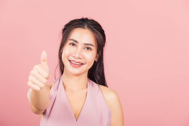 Portrait Asiatische schöne junge Frau lächelt sie stehend Finger Daumen nach oben, gutes Zeichen zuzustimmen Studioaufnahme isoliert auf rosa Hintergrund, thailändische Frau erfolgreich wie Fingergeste mit Kopierraum