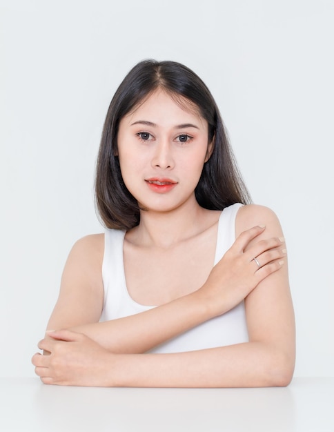 Portrait Beauty Shot, Nahaufnahme eines tausendjährigen asiatischen Modells mit kurzen schwarzen Haaren mit Make-up-roter Lippe im Tanktop-Unterhemd, das am Tisch sitzt und Blick in die Kamera auf weißem Hintergrund posiert.