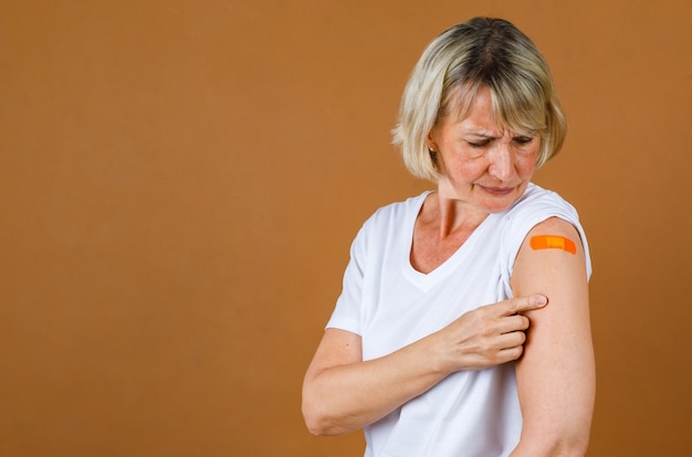 Portrait Closeup Studioaufnahme einer kaukasischen Seniorin blonder Patientin, die Stress betont, hat nach Erhalt der Covid-19-Impfung Schmerzen beim Blick auf die Injektionswunde an der orangefarbenen Gipsbinde auf ihrer Schulter.