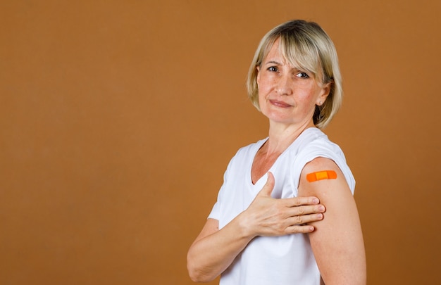 Portrait Closeup Studioaufnahme von kaukasischen älteren blonden Patientinnen, die betonen, dass sie Schmerzen mit einer Injektionswunde an einer orangefarbenen Gipsbinde auf ihrer Schulter haben, nachdem sie die Covid-19-Impfung erhalten haben.