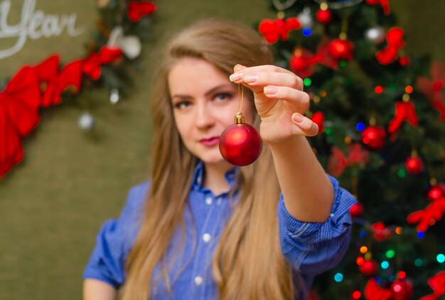 Portrait eines Mädchens mit leuchtend roten Lippen, blondes langes Haar Junges Mädchen in einem blauen Herrenhemd. eine Weihnachtskugel Dekoration vor sich halten. Ball essen