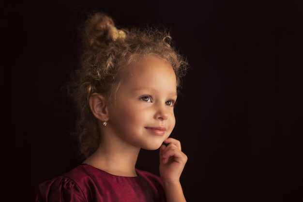 Portrait eines schönen kleinen Mädchens mit lockigem Haar und einer Schleife im burgunderfarbenen Kleid auf schwarzem Hintergrund