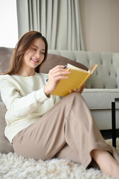 Portrait Entspannte asiatische Frau sitzt auf einem bequemen Teppich im Wohnzimmer und liest ein Buch