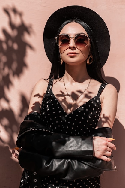 Portrait hübsche junge wunderschöne Frau in elegantem Hut in modischem Kleid in Vintage Lederjacke mit Sonnenbrille auf rosa Hintergrundwand am sonnigen Tag. Schönes Mädchen in Sommerkleidung genießen gute Laune.