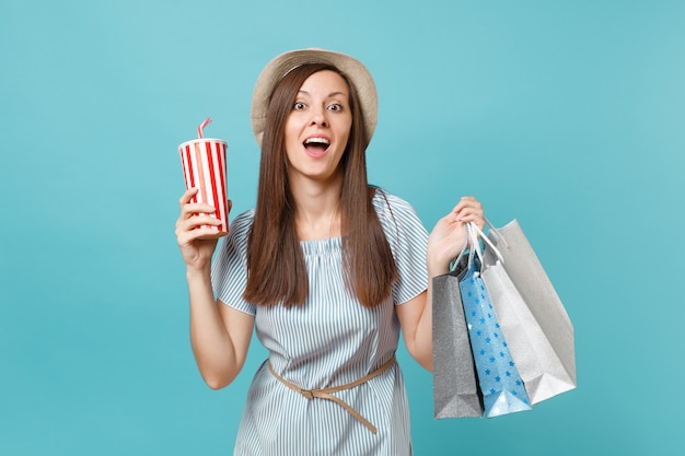 Portrait hübsches modisches lächelndes Mädchen im Sommerkleid, Strohhut, der Pakete mit Einkäufen nach dem Einkauf hält, Sodawasserbecher einzeln auf blauem Pastellhintergrund. Platz für Werbung kopieren