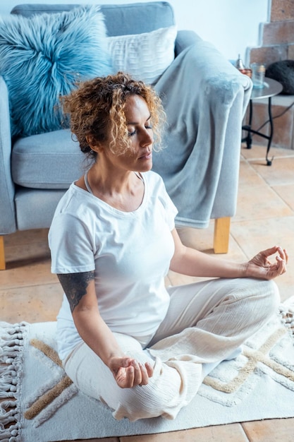 Portrait in voller Länge einer attraktiven jungen Frau, die zu Hause trainiert, Yoga-Übungen auf einer blauen Matte macht, in einer leichten, anständigen, angenehmen Haltung mit Palmen in Namaste sitzt, meditiert, atmet, entspannt