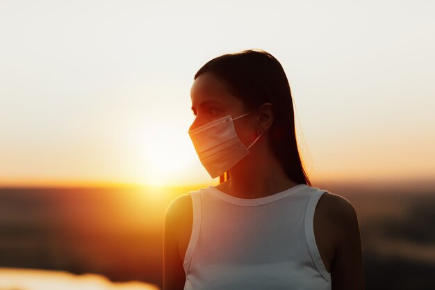 Portrait junge Frau, die medizinische Schutzmaske trägt, im Park am orange Sonnenuntergang während des Sommerabends stehend.