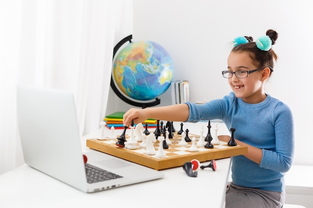 Portrait kaukasischen Little Cirly Mädchen, das Schach am Tisch im Innenraum mit Schachbrett spielt. kleines Mädchen lernt online Schach auf einem Laptop