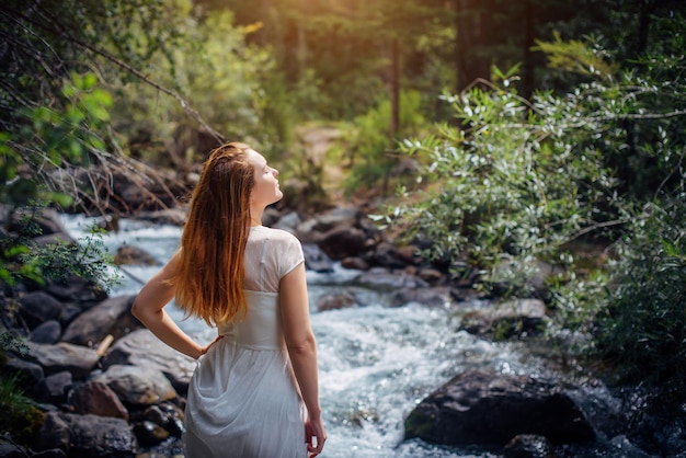 Portrait langhaarige Brünette in weißem Kleid posiert gegen kleinen Gebirgsfluss und grüne Bäume Schöne junge Frau am Ufer des Waldbachs