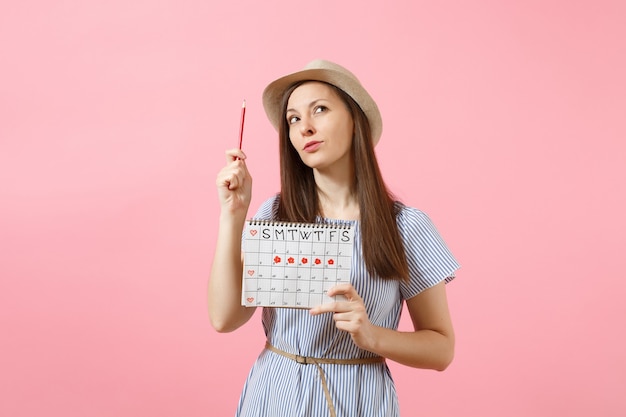 Portrait nachdenkliche Frau im blauen Kleid, Hut mit Rotstift, Kalender für weibliche Perioden zur Überprüfung der Menstruationstage einzeln auf rosafarbenem Hintergrund. Medizinisches Gesundheitswesen, gynäkologisches Konzept. Platz kopieren