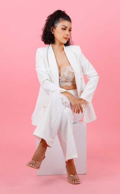 Portrait Nahaufnahme Studioaufnahme Asiatische junge sexy lockige Frisur professionelle erfolgreiche Geschäftsfrau im weißen Mode-Casual-Anzug und Spitzen-BH sitzend lächelnd gekreuzte Beine posiert auf rosa Hintergrund