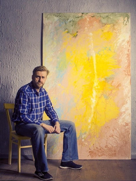 Foto portrait of male artist sitzt neben seinem abstrakten gemälde im studio