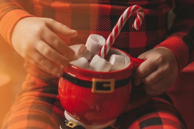 Portrait offen glückliches Kind im roten karierten Pyjama halten Weihnachtsbecher mit Marshmallows und Zuckerstange