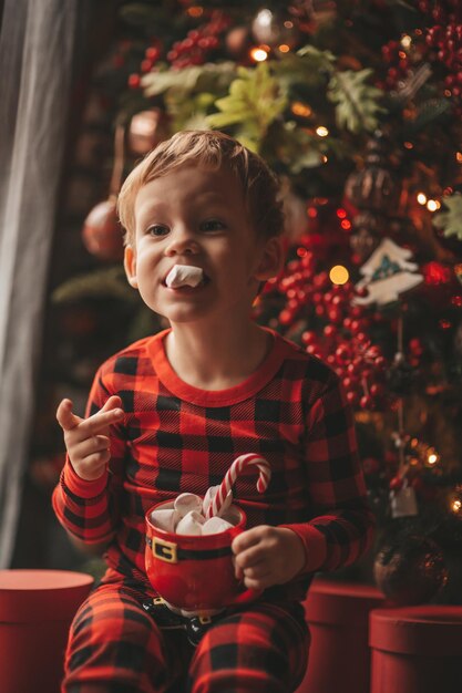 Portrait offen glückliches Kind im roten karierten Pyjama halten Weihnachtsbecher mit Marshmallows und Zuckerstange