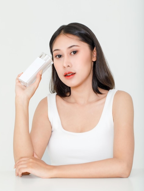 Portrait-Schönheitsaufnahme eines tausendjährigen jungen asiatischen, kurzen schwarzen Haares, weibliches Modell in Tanktop-Weste-Unterhemd, das ein Naturserum-Gesichtscreme-Hautpflege-Flaschenpaket in der Hand auf weißem Hintergrund hält.