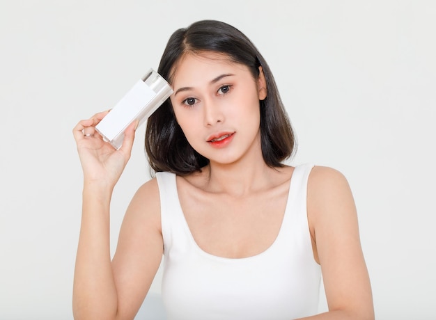 Portrait-Schönheitsaufnahme eines tausendjährigen jungen asiatischen, kurzen schwarzen Haares, weibliches Modell in Tanktop-Weste-Unterhemd, das ein Naturserum-Gesichtscreme-Hautpflege-Flaschenpaket in der Hand auf weißem Hintergrund hält.