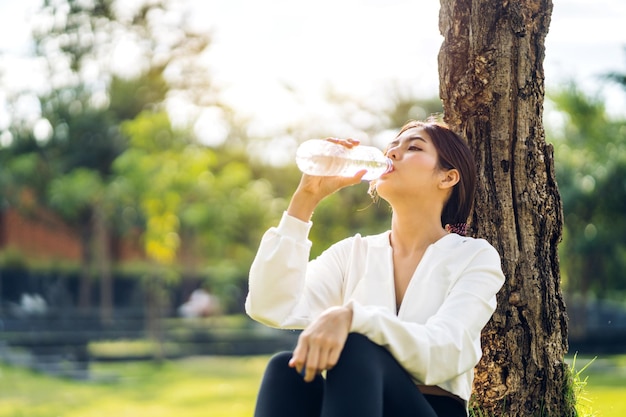 Portrait Sport asiatische Schönheit Körper schlanke Frau trinkt Wasser aus einer Flasche, während Sie sich entspannen und sich auf grünem Naturhintergrund im sommerlichen grünen Park frisch fühlenGesundes Lebensstilkonzept