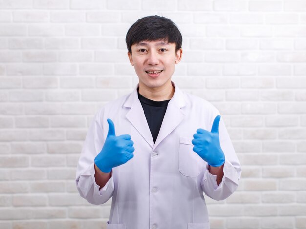 Portrait Studioaufnahme Asiatischer professioneller männlicher Wissenschaftler in weißen Laborkittel-Gummihandschuhen, der lächelnd in die Kamera auf Backsteinmauerhintergrund blickt.