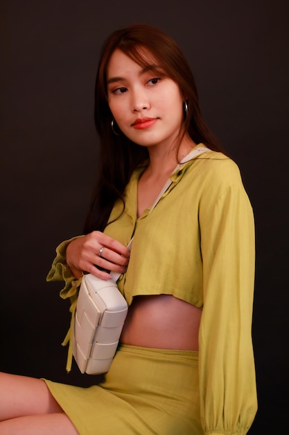 Portrait-Studioaufnahme einer asiatischen, urbanen Mode-Geschäftsfrau in lässigem Crop-Top-Outfit mit Handtasche, Blick in die Kamera auf schwarzem Hintergrund.